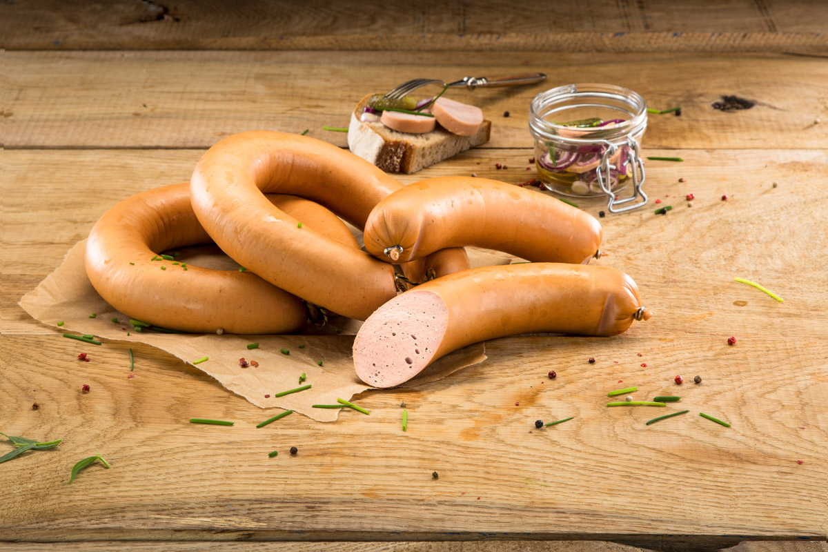 Mehrere Stücke Fleischwurst auf einem Holzbrett, dekoriert mit Kräutern und Pfefferkörnern, im Hintergrund ein Glas mit eingelegten Zwiebeln