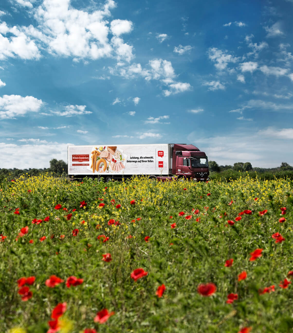 LKW von Wilhelm Brandenburg mit Werbebeschriftung für Fleischprodukte fährt durch ein Feld mit Blumen unter blauem Himmel