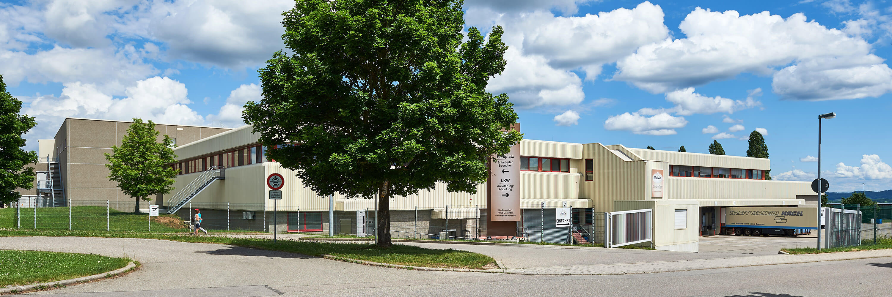 Industriegebäude von Wilhelm Brandenburg mit Bäumen im Vordergrund, umgeben von Zaun und Zufahrtsschildern bei blauem Himmel