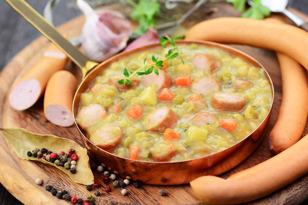 Erbsensuppe mit Wiener Würstchen, Karotten und Kartoffeln in einem kupferfarbenen Topf, umgeben von Gewürzen und frischen Zutaten