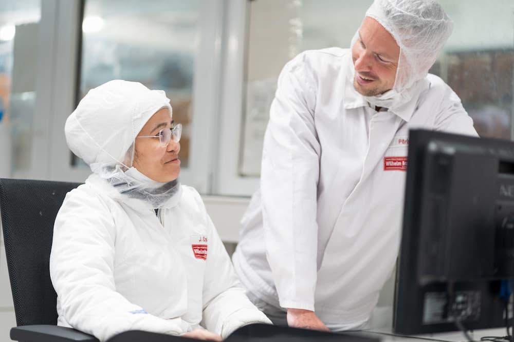 Zwei Mitarbeiter von Wilhelm Brandenburg in Schutzkleidung im Bürobereich, im Gespräch vor einem Computer