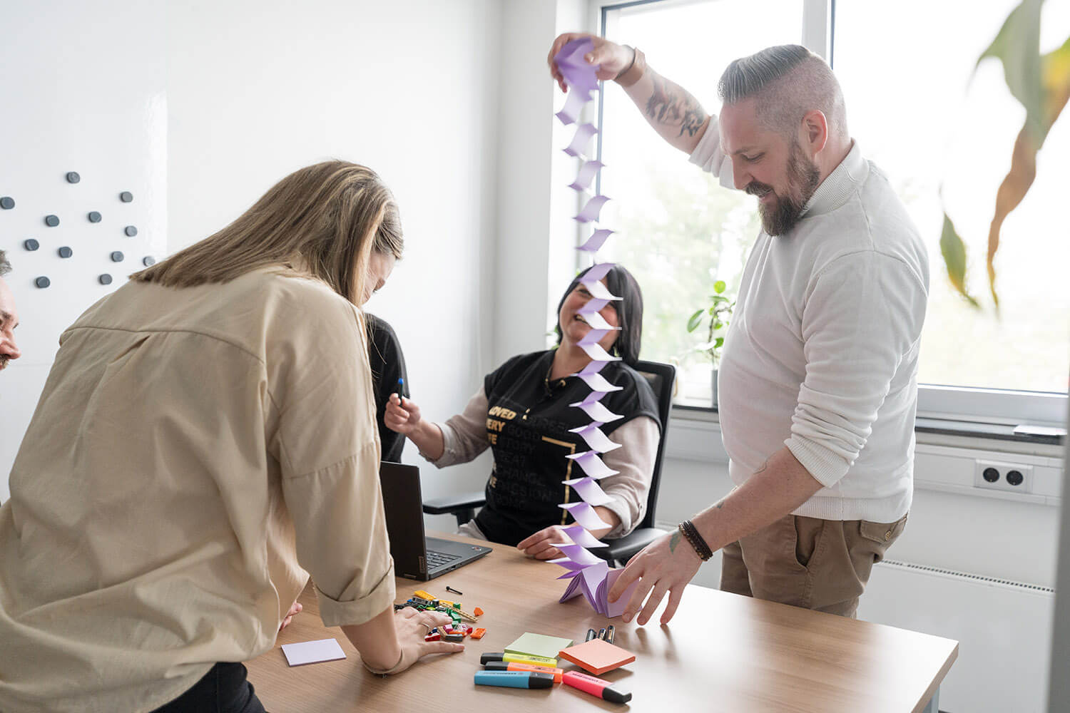 Kollegen arbeiten gemeinsam an einem Tisch, einer hält eine lila Papierkette hoch, während die anderen lachen und mit Materialien interagieren